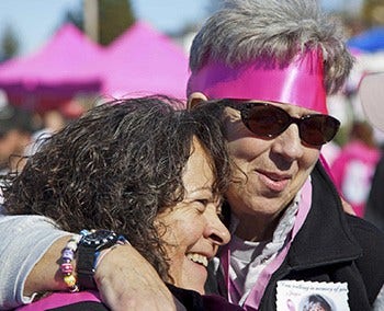 One woman with a pink bandanna is hugging another woman at a Strides Event outdoors.