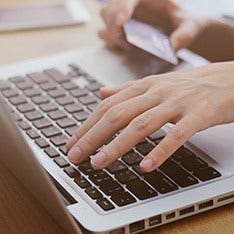 A person is typing on the laptop with his left hand while holding a credit card with his right hand in the background.